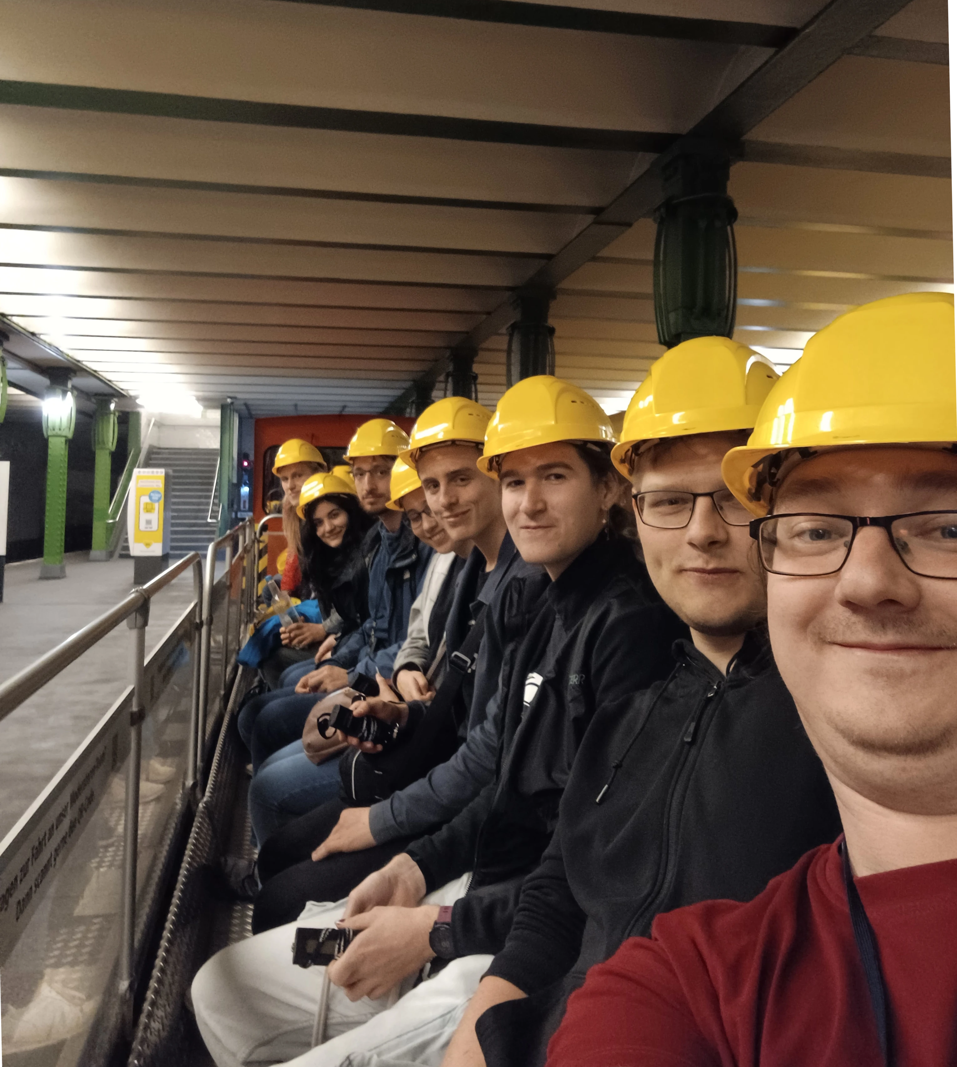 Mehrere Funkamateur:innen vom Funkclub DK0TU sitzen mit gelben Bau-Helmen auf einem offenen Bau-Wagen der Berliner U-Bahn und schauen glücklich in die Selfi-Kamera