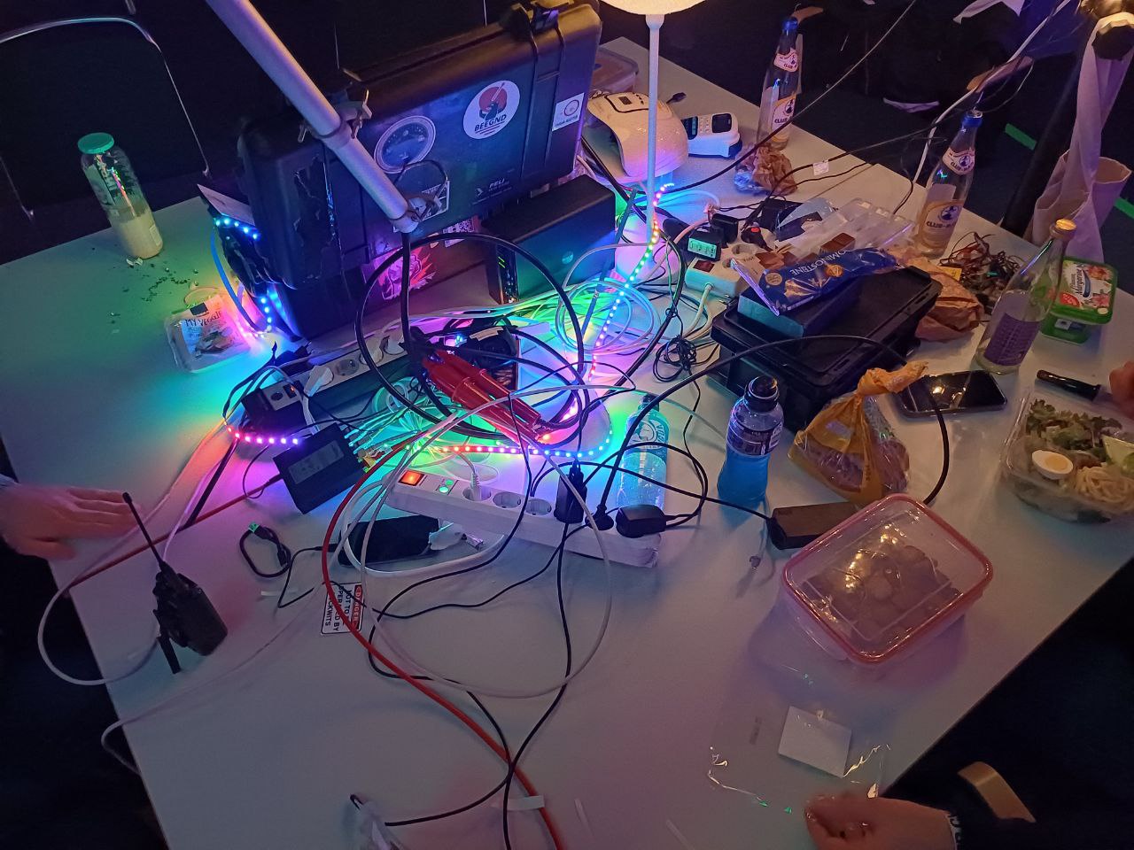 Tisch, mit leuchtendem LED-Streifen, einem schwarzen Flight-Case, vielen Kabeln, etwas Toastbrot, Aufstrich und einigen Mate-Flaschen